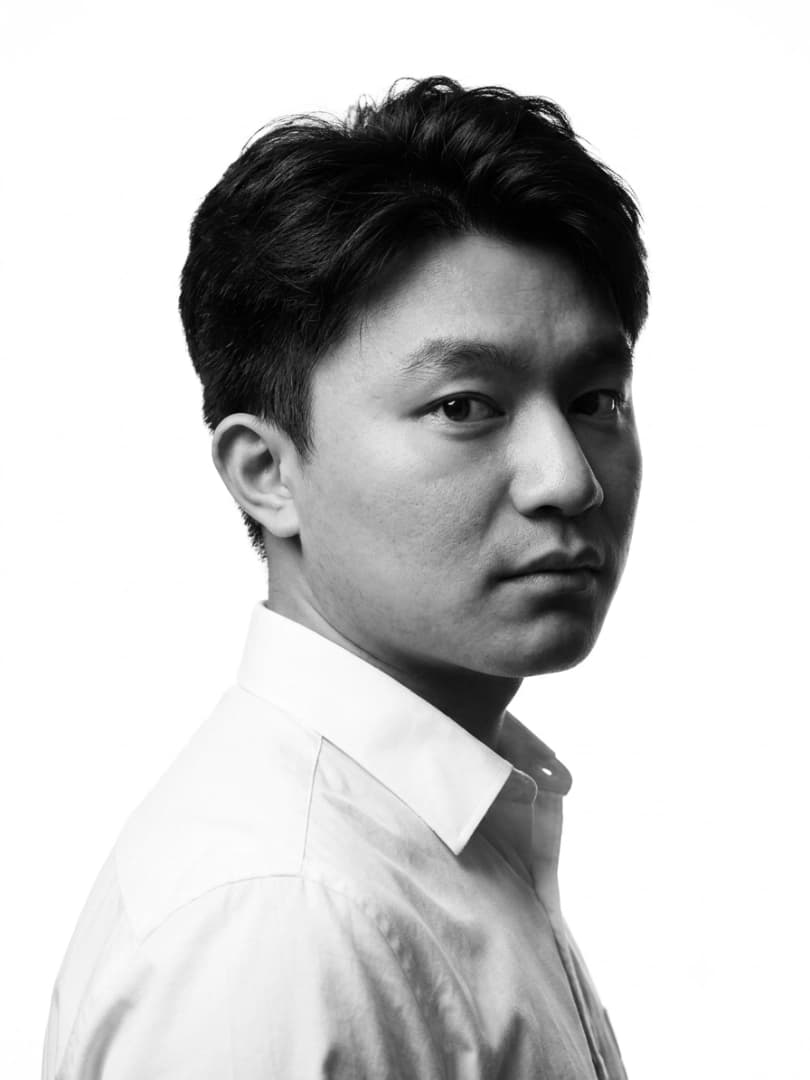 Ngawang Chojey Rai, shoulder to the camera with his head turned back to the lens, in a white shirt against a plain ground.